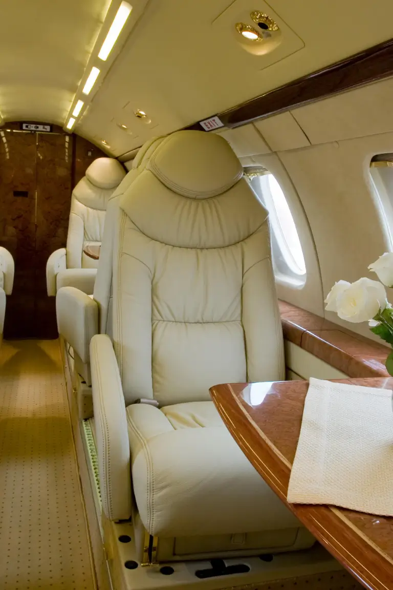 a luxurious and comfortable white leather seat inside a private jet with a polished wooden table and a white napkin on it with additional seats an EXIT sign and two windows in the background