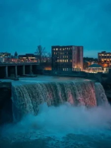 A scenic view of a magnificent high falls in Rochester New York illuminated under the twilight sky, with city buildings and lights in the background