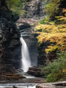 breathtaking view of Watkins Glen State Park in Rochester, NY, showcasing a serene waterfall cascading down rugged cliffs, surrounded by lush autumn foliage and an ancient stone bridge arching over the gorge.